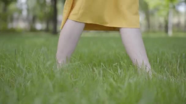 Gambe femminili in abito giallo a piedi nudi a piedi nudi gettare l'erba verde nel parco in tempo ventoso primo piano . — Video Stock