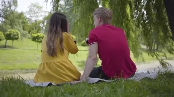 Joven pareja feliz en el amor haciendo picnic con vino en el hermoso jardín floreciente o parque charlando alegremente y sonriendo. Fecha de joven hermoso hombre y mujer. Romántico pasar tiempo al aire libre . — Vídeos de Stock