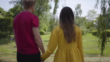 Güzel gölün yakınındaki yeşil parkta yürüyen sevimli mutlu çift, bahar güzel gününde birbirlerinin elini hafifçe tutuyor. Romantik vakit geçirmek dışarıda.