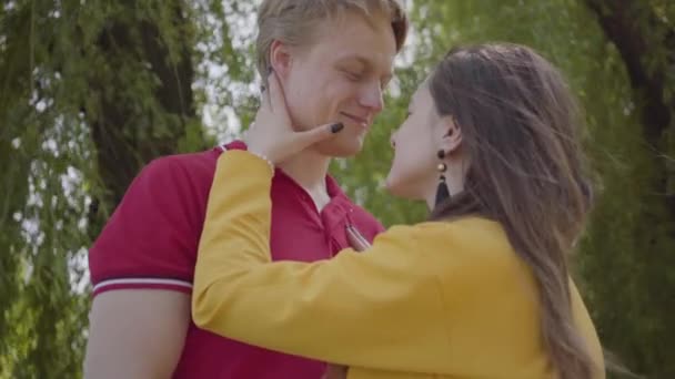 愛らしい白人のカップルは、緑の春の公園の背景に優しく抱きしめ、微笑んでいます。若い美しい男女の日付。屋外でロマンチックな時間を過ごす. — ストック動画