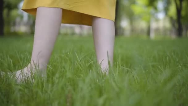 Las piernas femeninas en vestido amarillo caminando descalzas arrojan la hierba verde en el parque en un clima ventoso de cerca. Movimiento lento . — Vídeos de Stock