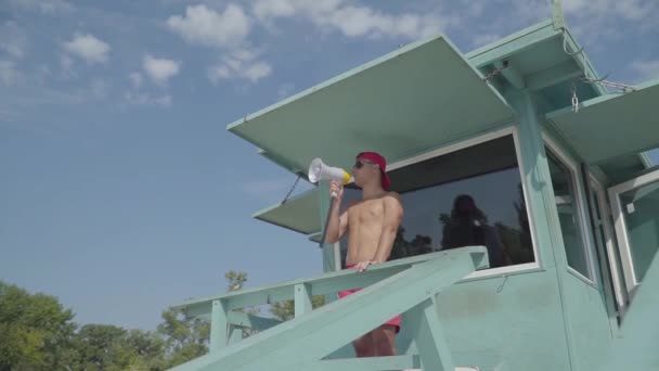 Habilidad sin camisa salvavidas atlético en gafas que hablan en una situación de control de gritos en la playa. El hombre mirando a los bañistas por turistas. Concepto de profesión — Vídeo de stock