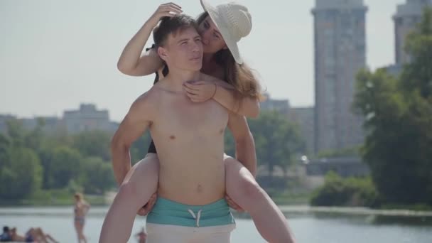 Feliz pareja joven divirtiéndose en la playa. La chica sentada en la espalda de un tipo sonriendo. Los amantes de disfrutar de los fines de semana cerca del agua, río en el día de verano soleado caliente. Pasatiempos de verano ocio actividades concepto — Vídeos de Stock