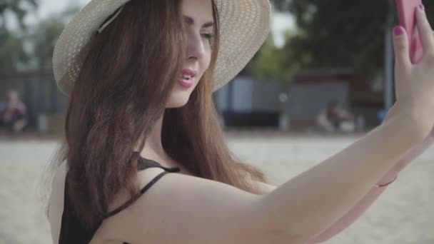 夏の白い帽子をかぶった若い女性の肖像画は、ビーチで休んで携帯電話で自分撮りを取ります。夏のレジャーコンセプト。週末の時間 — ストック動画