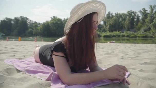 Retrato de mujer joven en el sombrero blanco de verano acostado en la playa. Concepto de ocio de verano. Hora del fin de semana. Hermoso paisaje en el fondo — Vídeo de stock