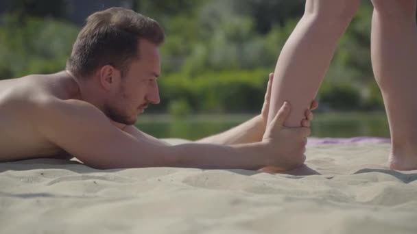 Привлекательный мужчина лежит на песке, держа за ногу молодую женщину, которая пытается ходить крупным планом. Обучение на открытом воздухе. Активный образ жизни. Время для спорта — стоковое видео
