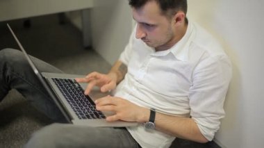 Ofiste yerde oturan ve duvara yaslanan adam. Adamın elinde dizüstü bilgisayarı var ve daktilo ediyor..