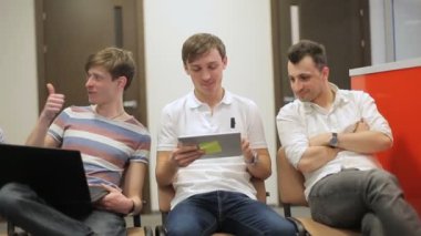 Üç adam sandalyede oturuyor ve konuşuyorlar. Adam gülümsüyor. İkinci adam tablet üzerinde çalışıyor. Üçüncü adam gülümsüyor ve elinde dizüstü bilgisayar var. Kızlar konuşuyor..