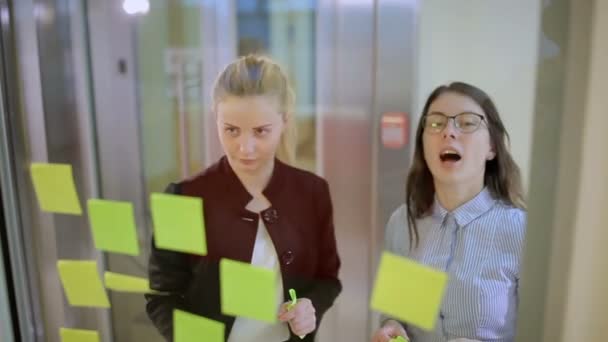 Две Молодые Девушки Обсуждают Проблему Офисе Блондинка Приклеивает Наклейки Стекло — стоковое видео