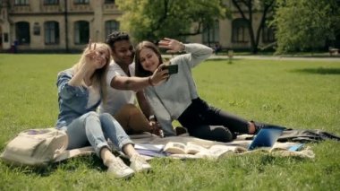 Kampüs bahçesinde battaniye üzerinde oturan ve telefonda selfie yapmak ve merhaba, video sallayarak üç çekici öğrenciler  