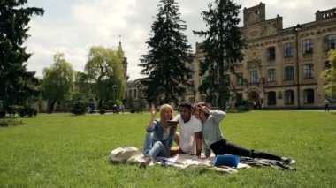 Parkta çim üzerinde battaniye üzerinde oturan çekici çok ırklı öğrenciler, Üniversite binası ile arka plan, video  