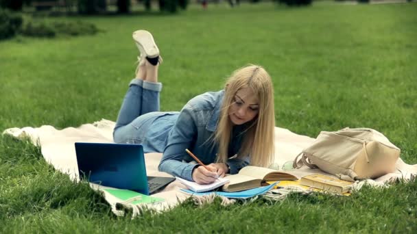 屋外で鉛筆を手にしたクローズアップ美しい学生読書本 長髪のブロンドの学生は 勉強中に公園のキャンパスの芝生の毛布の上に横たわって ビデオ — ストック動画