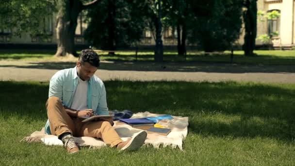 ハンサムなアフロアメリカ人学生が公園で毛布の上に座って勉強しています 男性学生はタブレット上でページをスクロールし ヘッドフォンを介して音楽を聞きながらノートブック上でいくつかのノートを作成しています — ストック動画