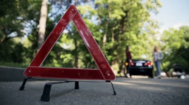 Ön taraf kırmızı acil durum üçgenine yakın, arka planda iki çekici kadın bozuk bir arabanın yanında duruyor. Araba yol kenarına park edilmiş ve acil durum dur sinyali yanıyor..