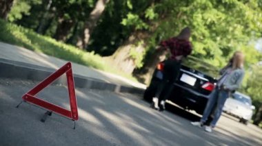 Öndeki kırmızı acil durum kırmızı dur işareti yolda, arka planda bulanık genç kadınlar kırık arabanın önünde duruyor ve acil durum sinyali yanıyor..