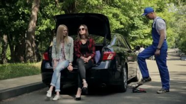 Genç kadınlar açık bagajda oturuyorlar. Araba yol kenarına park edilmiş, çünkü lastiği patlamış. Tamirci araba lastiklerini şişiriyor. Kızlar sohbet edip dinlenirken ayak pompası kullanıyor.