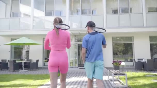 Pohled na mladého páru s raketama jde do hotelu. Dívka v růžového oblečení a muž v modrém sportovním oblečení. Aktivní volný čas. — Stock video