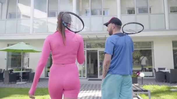 Widok z tyłu młodej pary z rakietami udać się do hotelu. Dziewczynka w ubraniach różowych i mężczyźnie w niebieskiej odzieży sportowej. Aktywny wypoczynek. — Wideo stockowe
