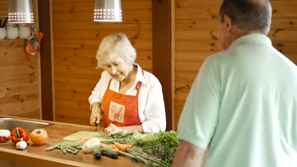 年配の妻に花を贈る男キッチンに立っている成熟した女性 野菜をカット ビデオ — ストック動画