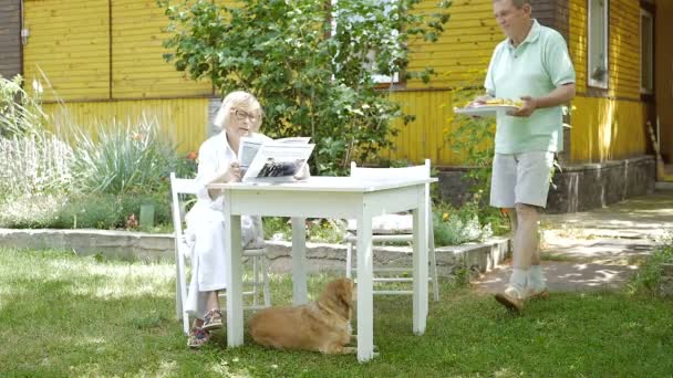 庭のタブレットに座っている高齢の女性 食べ物のトレイを持って来る男 ビデオ — ストック動画