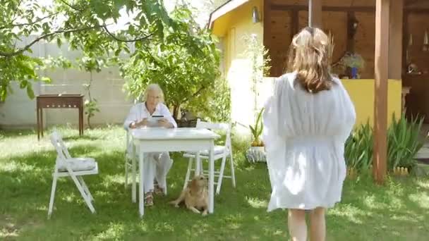 一位老妇人坐在花园里 手里拿着一块石碑 一个年轻的女人带着一个盘子 带着午餐在花园里吃 女儿给她母亲举了一个太阳镜的例子 — 图库视频影像