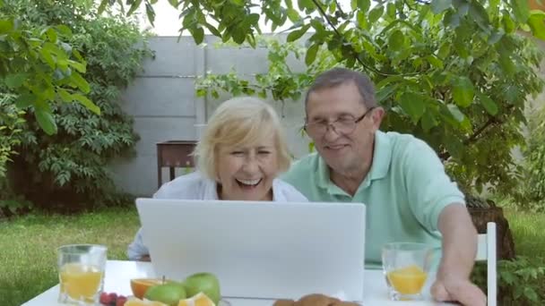 庭でノートパソコンを通して話している老夫婦 庭にノートパソコンを持って座っている老夫婦 庭の白いテーブルの上に立つ果物とジュースのプレート — ストック動画