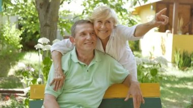 Kadın bankta oturmuş kocasına sarılıyor. Erkek ve kadın bahçede oturuyor ve gülümsüyor. Kadın kocasına bankta oturduğunu söylüyor..