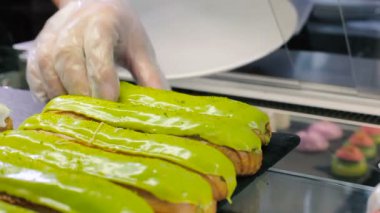 Ele tutulur. Bir kadın tabağa iki ekkin koyar. Yeşil kremayla süslenmiş ekler. Şekerci, selofan eldivenlerle çalışır. Satıcı bir tabağa iki ekler ve dükkanın camını kapatır. Yakın plan..