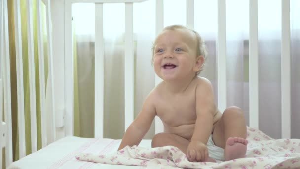 婴儿坐在婴儿床上笑着 小女孩坐在一个婴儿床里 婴儿看了看旁边 穿尿布 手拿着尿布的女孩 — 图库视频影像