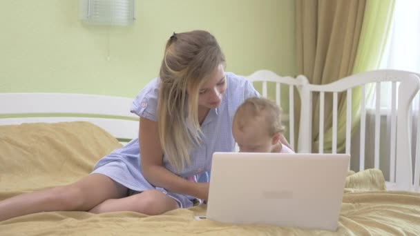 ノートパソコンのキーボードを入力する赤ちゃん 小さな女の子が開き ノートパソコンの蓋を閉じます 白いラップトップの近くのベッドに座っている赤ちゃんを持つお母さん ピンクのドレスを着た赤ん坊と頭のための包帯 — ストック動画