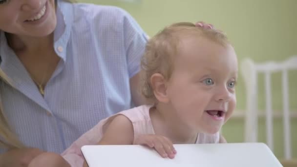 婴儿移动笔记本电脑的盖子 妈妈和宝宝在镜头前挥手 妈妈抱着孩子坐在白色笔记本电脑旁边的床上 穿着粉色连衣裙 头戴绷带的婴儿 — 图库视频影像
