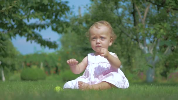 庭の草の上に座っている少女 少女はりんごを拾い上げ 口に持ってくる 少女は手にりんごを考えている ドレスを着た女の子 — ストック動画