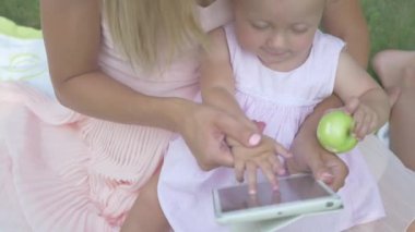 Anne kızını tablet ekranın elinden tutuyor. Anne ve kızı tablete bakıyor. Annesi bahçede oturan bir bebek. Küçük kız yeşil elmayı ısırıyor..
