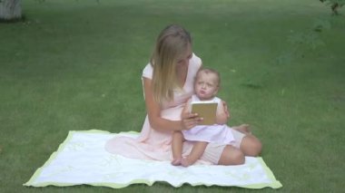 Bebeği battaniyenin üzerinde oturan bir kadın. Anne kızını tablet ekranın elinden tutuyor. Anne ve kızı tablete bakıyor. Annesi bahçede oturan bir bebek. Küçük kız yeşil elmayı ısırıyor..