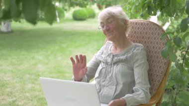 Laptop kameralı bir kadın. Laptopta konuşan yaşlı bir kadın. Sallanan sandalyede oturan yaşlı kadın. Dizlerinin üzerinde beyaz bir dizüstü bilgisayar tutan bir kadın. Bir kadın bahçede bir ağacın altında dinleniyor..