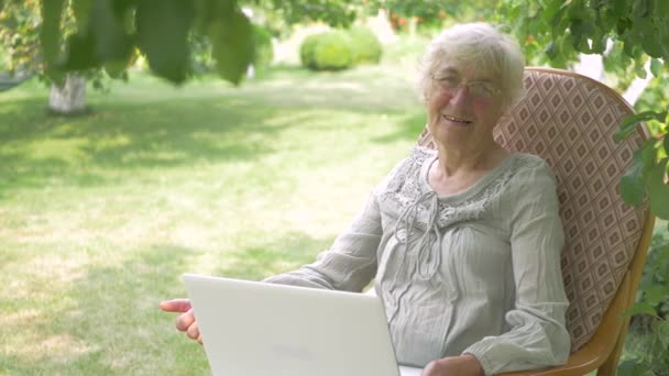 カメラに親指を立てた年配の女性 高齢の女性はロッキングチェアに座っている 膝の上の女性は白いノートパソコンを持っている 庭の木の下に女性が休んでいる — ストック動画