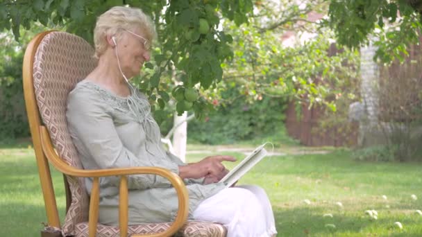 老年妇女打开平板电脑上的音乐 奶奶坐在椅子上跳舞 女人在耳机里听音乐 戴眼镜的女人躺在花园里 老妇人坐在椅子上 — 图库视频影像
