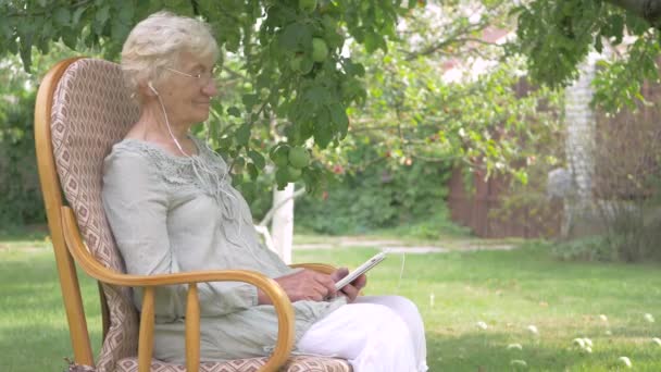 女人在椅子上荡秋千 听音乐 女人在耳机里听音乐 戴眼镜的女人躺在花园里 老妇人坐在椅子上 — 图库视频影像