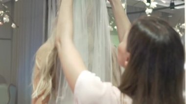 Yakın plan: kızın kafasına duvağı takan asistan. Asistan peçeyi düzeltti ve onu kızın omuzlarına koydu. Kamera aynadaki yansımaya odaklanıyor. Gelin gülümsüyor..