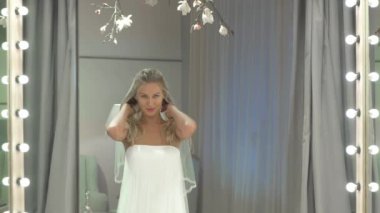 Kamera, gelinin aynadaki yansımasına odaklanıyor. Kız gülümsüyor ve aynada kendine bakıyor. Gelin saça dokunuyor ve duvağı düzeltiyor. Gelin kendine farklı yönlerle bakıyor..