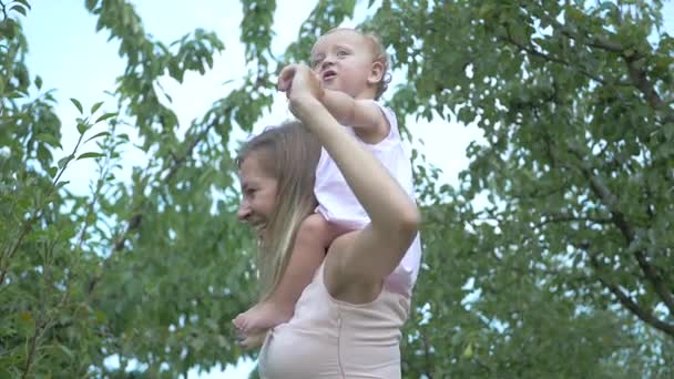 宝宝坐在妈妈的脖子上 妈妈把她的女儿 女儿和她的母亲在花园里散步 女人跳着抱着一个孩子 穿着粉红衣服和尿布的婴儿 慢动作 — 图库视频影像