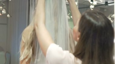 Yakın plan: kızın kafasına duvağı takan asistan. Asistan peçeyi düzeltti ve onu kızın omuzlarına koydu. Kamera aynadaki yansımaya odaklanıyor. Gelin gülümsüyor..