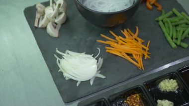 Yakın plan. Kamera soldan sağa hareket ediyor. Kara tahtada dilimlenmiş soğan, havuç, kuşkonmaz ve mantar var. Ayrıca masada dilimlenmiş et ve diğer malzemeler de var..