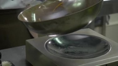 Yakın plan. Aşçı sıcak bir tavada sebzeleri kepçeyle karıştırıyor. Adam eline bir tava alır ve pişmiş sebzeleri kepçeyle süzgece koyar. Aşçı elinde yağ olan sarı bir şişe alır..