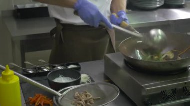 Yakın plan. Aşçı sıcak tavada havuç ve mantarı kepçeyle karıştırıyor. Adamın eli masadan bir kuşkonmaz alıp tavaya koydu. Aşçı karıştırmaya devam ediyor. Aşçı sebzeli bir tavaya biber koyar ve yemeği karıştırır..