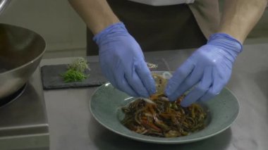 Yakın plan. Sebze ve diğer farklı malzemelerle dolu tabak mutfaktaki masanın üzerinde duruyor. Aşçının elleri eldivenle tabağı limonla süslüyor ve susam tohumlarını bir tabağa ekliyor. Adam tabağa biraz yeşil baharat koydu..