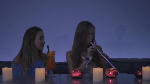 两个漂亮的女孩坐在咖啡厅里重拍 一个长头发的可爱女孩抽着妓女 小女孩冒着浓烟 微笑着 对她的朋友说几句话 然后把烟斗递给她的朋友 — 图库视频影像