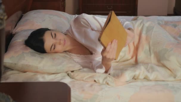 年轻女人躺在床上 手里拿着一块石碑 女人用平板电脑 关上床罩 把小玩意放在床边的木制桌子上 年轻的女人闭上眼睛睡着了 — 图库视频影像
