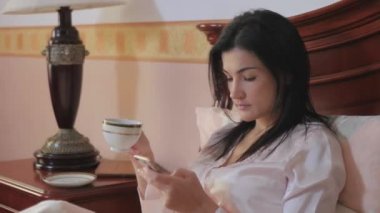 Genç esmer kadın yatakta oturuyor ve sağ elinde çay, sol elinde akıllı telefonu olan bir fincan tutuyor. Genç esmer kadın telefonunun ekranına bakıyor ve fincandan çay içiyor..