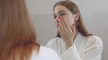 Farklı renkli gözleri ile çekici genç kadın banyoda yüzüne günlük krem uygulayarak aynaya bakıyor. Cilt bakımı, güzellik konsepti. Gerçek insanlar serisi.